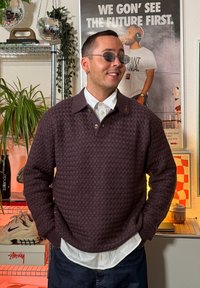 Bordowy sweter z dzianiny z fakturowanym wzorem, kołnierzykiem i guzikiem, noszony na białej koszuli. Okrągłe okulary przeciwsłoneczne i casualowe ciemne dżinsy.