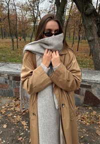 Femeie îmbrăcată într-un palton bej și o eșarfă gri care își acoperă gura, stând într-un parc de toamnă cu frunze căzute și copaci dezgoliți.