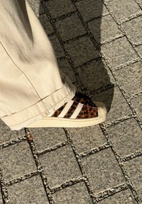 Sapatilhas com padrão de leopardo e riscas creme, parte superior texturizada e sola de borracha bege. Usadas com calças largas de cor creme sobre um pavimento de pedra.