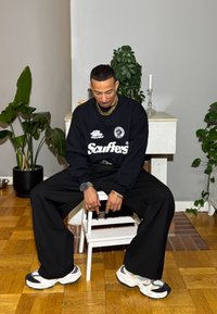 Fekete pulóver "Scuffers" felirattal, laza fekete nadrág, vastag fehér és fekete sportcipők, arany kiegészítők; fehér széken ülve.