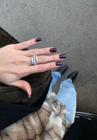 Anel triplo em prata com pedras claras, usado numa mão com unhas de um roxo escuro; a mão repousa sobre uma bolsa preta texturizada.