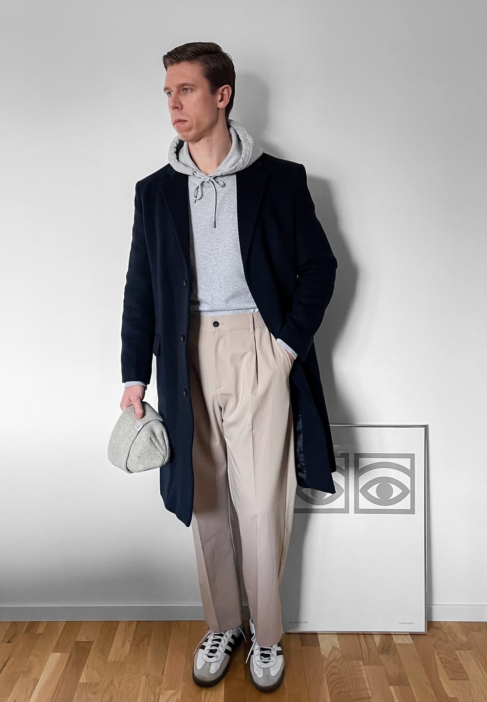 Homme portant un manteau bleu marine, un sweat à capuche gris, un pantalon beige et des baskets, tenant une casquette grise, debout près d'une œuvre d'art abstraite représentant un œil encadrée sur le sol.