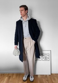 Homme portant un manteau bleu marine, un sweat à capuche gris, un pantalon beige et des baskets, tenant une casquette grise, debout près d'une œuvre d'art abstraite représentant un œil encadrée sur le sol.