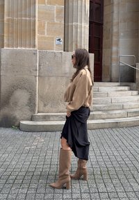 Beżowy, krótki sweatshirt, czarna asymetryczna spódnica i beżowe botki na wysokim obcasie do kolan. Widok z profilu obok kamiennej ściany i schodów.