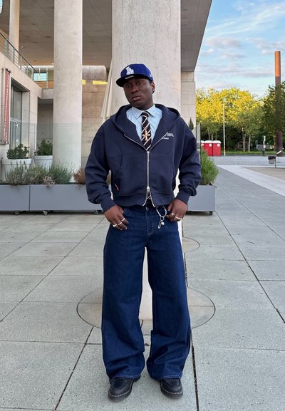 Sudadera con cremallera navy, camisa de cuello azul claro, corbata con estampado, pantalones de mezclilla anchos azules, botas negras, acentos de joyería, con una gorra de béisbol.