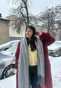 Cappotto rosso e soffice sopra un maglione giallo, sciarpa grigia con frange. Berretto di lana nero, in piedi in un ambiente esterno innevato con delle automobili.