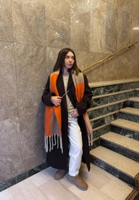 Femme en manteau noir, écharpe orange et beige, pantalon blanc et bottes marron clair, debout près d'un escalier en marbre avec une rampe en laiton à l'intérieur.