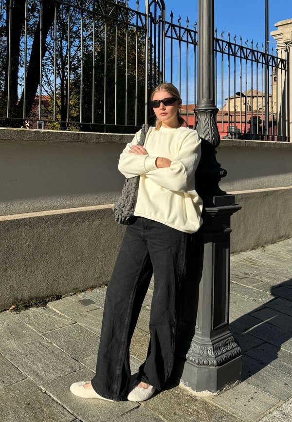 Sudadera de gran tamaño color crema, pantalones de pierna ancha negros y zapatillas blancas de felpa, con un bolso texturizado gris. De pie junto a una farola.