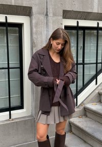 Hnědý trenčkot s páskem přes tmavý svetr, v kombinaci s plisovanou �šedou sukní a hnědými kozačkami po kolena, stojící na kamenných schodech.