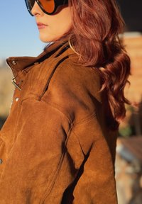 Giacca in camoscio marrone con finitura testurizzata, dotata di chiusura a zip e dettagli sulle spalle, abbinata a grandi occhiali da sole scuri.