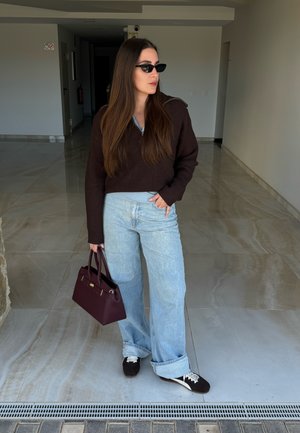 Mujer con el cabello largo y castaño, lleva gafas de sol oscuras, un suéter marrón, jeans anchos de color azul claro, zapatillas negras y sostiene un bolso de mano de color púrpura oscuro.