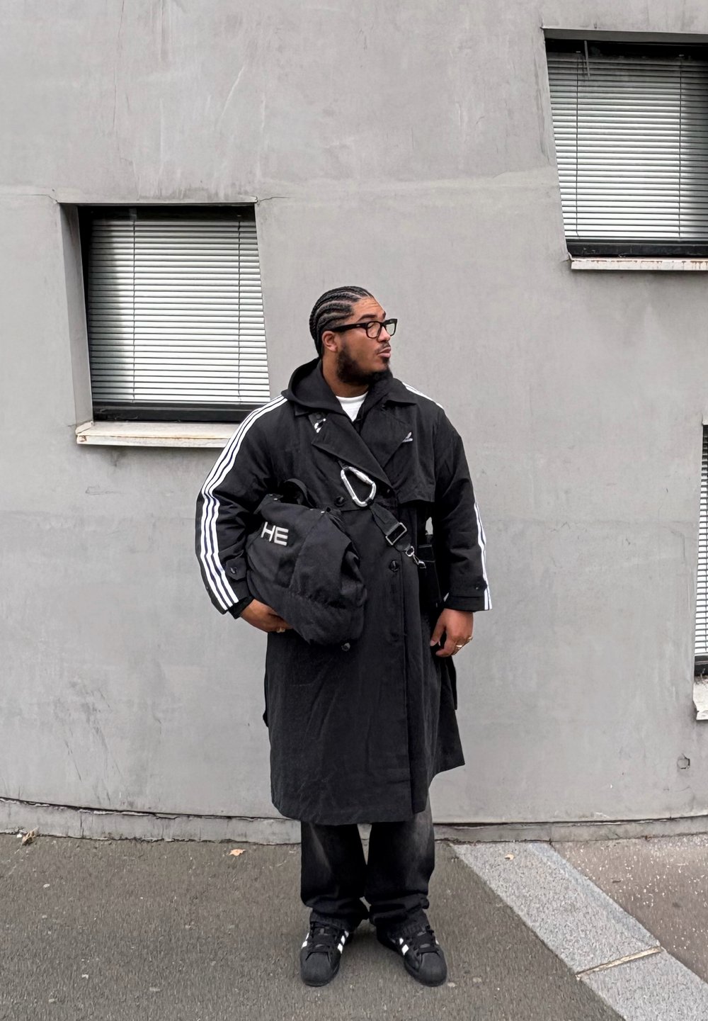 Long manteau noir avec des rayures blanches, sac noir avec détails métalliques, pantalon noir et baskets. En arrière-plan, un mur en béton et des fenêtres.