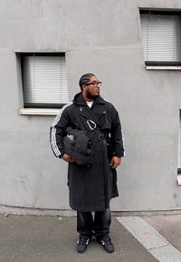 Long manteau noir avec des rayures blanches, sac noir avec détails métalliques, pantalon noir et baskets. En arrière-plan, un mur en béton et des fenêtres.