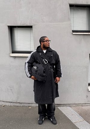 Manteau long noir avec des rayures blanches, sac noir avec détails métalliques, pantalon noir et baskets. En arrière-plan, un mur en béton et des fenêtres.