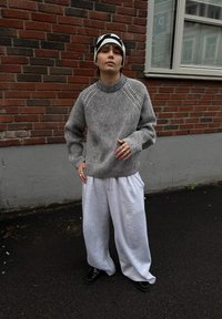 Suéter de rayas gris y negro tejido con cuello acanalado, combinado con pantalones deportivos blancos holgados. Zapatos negros y un gorro estampado completan el conjunto.