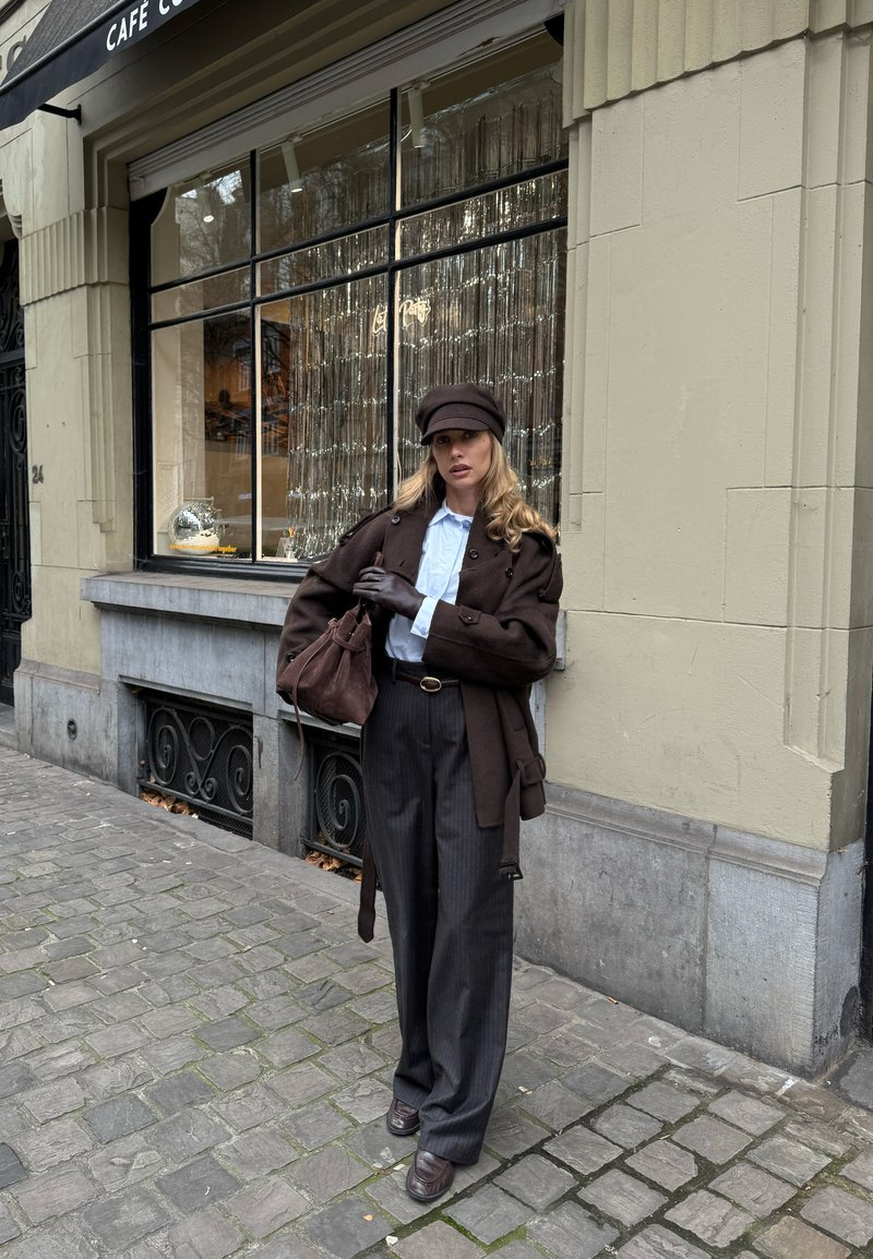 Brauner Wollmantel über einem hellblauen Hemd, Nadelstreifenhose und einer braunen Mütze. Hält eine braune Tasche, steht auf Kopfsteinpflaster vor einem Geschäft.