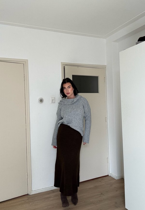 Kvinde med mørkt hår iført en grå sweater, lang mørk nederdel og mørke støvler, der står indendørs nær lukkede beige døre.