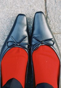 Svarta spetsiga ballerinaskor med små rosetter, över ljusröda strumpor, på ett grått kakelgolv.