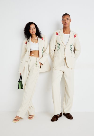 Blazers beige con flores rojas bordadas y diseños de serpiente verde, combinados con pantalones a juego y un bolso negro. Sandalias de tiras y zapatos de estampado animal.