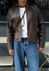 Person som bär en öppnad mörkbrun läderjacka, vit t-shirt, blå jeans och ett brunt diagonalaxelband över bröstet.