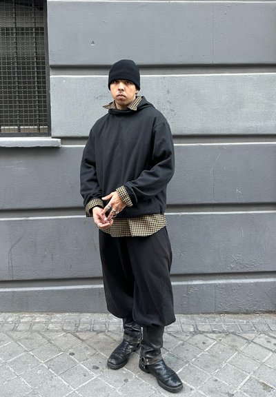Sudadera negra oversized superpuesta sobre una camisa a cuadros en tonos marrones, combinada con pantalones negros holgados y botas negras hasta la rodilla con hebillas.