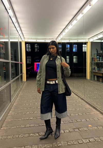 Chaqueta verde sobre una blusa negra, culottes de mezclilla anchos con puños de mezclilla y botines negros de punta. Bolso negro y joyería dorada.