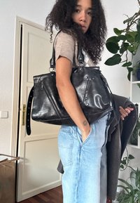 Žena s kudrnatými vlasy, oblečená v béžovém topu a světlých džínách, nese velkou černou koženou tašku a má v ruce šedou bundu uvnitř budovy.