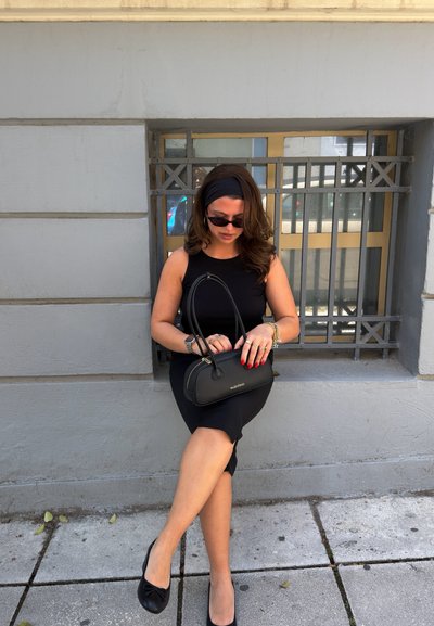 Mujer con vestido negro y banda en la cabeza sentada en un borde junto a una ventana, sosteniendo un bolso Valentino negro y llevando gafas de sol.