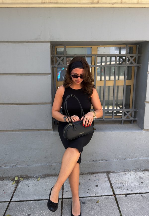 Femme en robe noire et bandeau assise sur un rebord près d'une fenêtre, tenant un sac à main Valentino noir et portant des lunettes de soleil.