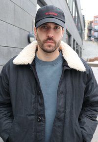 Homme avec barbe et boucles d'oreilles portant une casquette noire, une veste noire avec un col en polaire blanche, et un sweat-shirt bleu debout près d'un bâtiment gris.