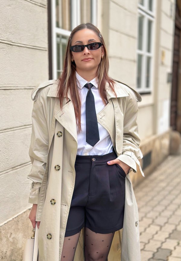 Vrouw met lang haar die een zonnebril, beige trenchcoat, wit hemd, zwarte das, zwarte short en een stippenpanty draagt, staand op straat.