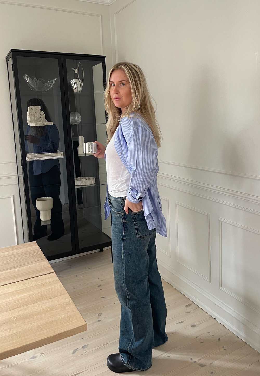 Una donna si trova accanto a una vetrina in vetro, indossando una camicia a righe blu e jeans blu scuri, mentre tiene in mano una coppa d'argento. Un tavolo in legno chiaro è nelle vicinanze.