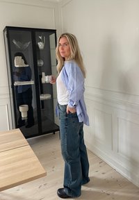 Una donna si trova accanto a una vetrina in vetro, indossando una camicia a righe blu e jeans blu scuri, mentre tiene in mano una coppa d'argento. Un tavolo in legno chiaro è nelle vicinanze.