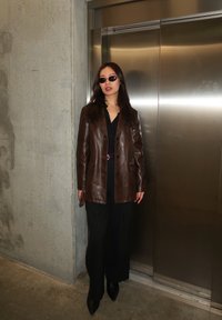 Femme portant des lunettes de soleil, une veste en cuir marron, une tenue noire, debout près de portes d'ascenseur métalliques fermées dans un espace intérieur aux murs en béton.