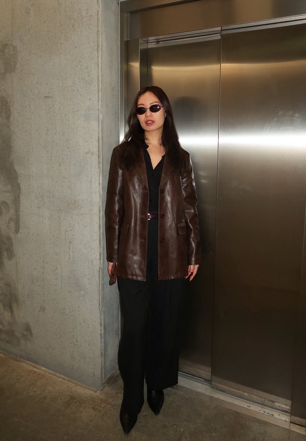 Femeie purtând ochelari de soare, jachetă din piele maro, ținută neagră, stând lângă uși metalice închise de lift într-o zonă cu pereți din beton, în interior.