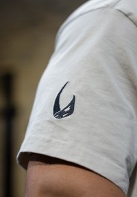 Lys grå kortermet skjorte med en mørkeblå brodert logo på venstre erme. Myk stoff med en jevn tekstur.