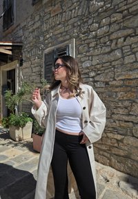 Manteau long beige avec de grands boutons, associé à un débardeur blanc et à un pantalon noir. Texture lisse, coupe décontractée et cadre extérieur visible.