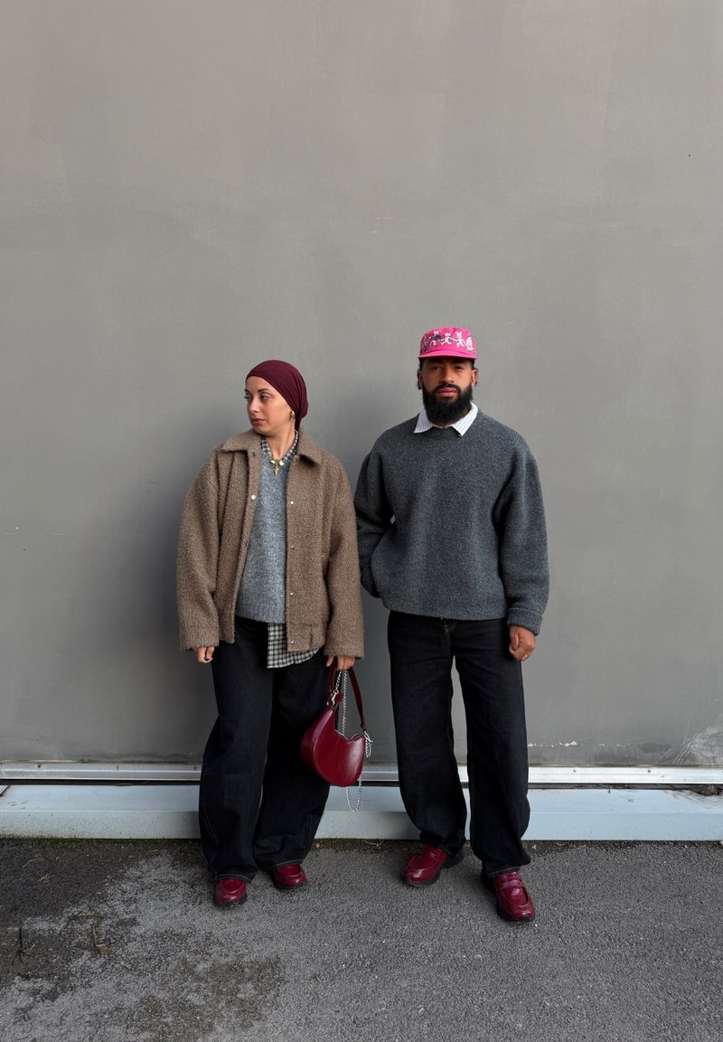 Deux personnes sont debout contre un mur gris ; toutes deux portent un pantalon foncé et des chaussures bordeaux, l'une un blouson marron et un foulard, l'autre un pull gris et une casquette rose.