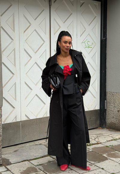 Mujer con cabello trenzado lleva un abrigo largo negro, atuendo negro con un detalle floral rojo, sosteniendo un bolso de mano negro, de pie en una acera.