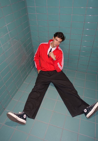 Chaqueta roja con cremallera y rayas blancas, pantalones negros de pierna ancha y zapatillas negras de caña alta. Fondo de pared de azulejos color verde azulado.