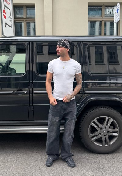 Una persona vestida con una camiseta blanca ajustada y vaqueros negros holgados está de pie al lado de un SUV Mercedes-Benz G-Class negro, que destaca por sus acentos plateados.