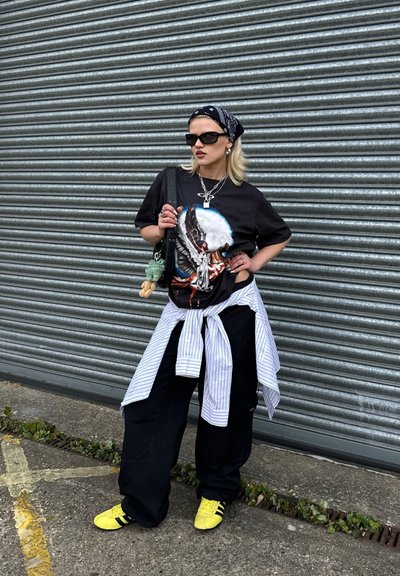 Camiseta gráfica negra con un estampado de un ángel. Combinada con pantalones cargo negros y zapatillas Adidas amarillas. Accesorios de cadenas y un pañuelo.