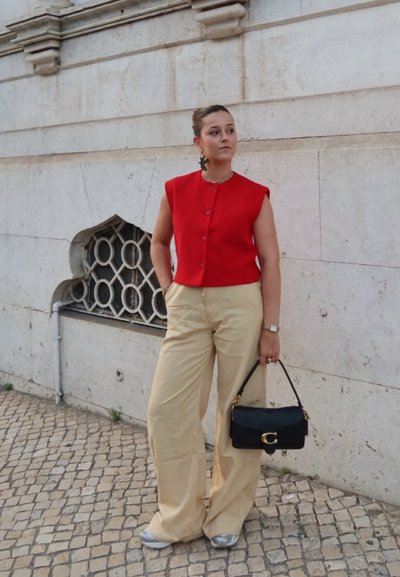 Top sin mangas rojo con botones, pantalones de pierna ancha beige, bolso negro con detalles dorados, combinado con zapatillas, contra una pared texturizada.