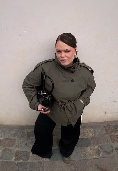 Mujer con cabello oscuro con una chaqueta verde oliva y pantalones negros sosteniendo un bolso negro contra una pared beige en una calle empedrada.