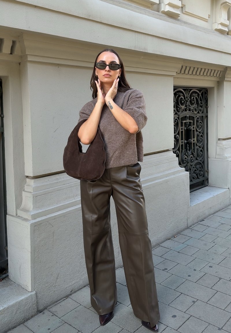 Femme portant des lunettes de soleil foncées, un pull marron, un pantalon en cuir, et tenant un sac à main marron foncé, debout sur un trottoir près d'un bâtiment clair.