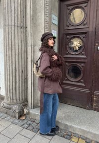 Veste marron surdimensionnée avec texture duveteuse et ceinture, associée à un jean bleu à jambes larges et un sac bandoulière à motif. Des bottes noires complètent la tenue.