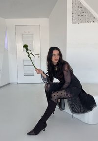 Schwarzes, durchsichtiges Kleid mit langen Ärmeln und Punkten-Strumpfhosen. Stöckelschuhe in dunkler Farbe, hält eine grüne Blume, minimalistischer Hintergrund.
