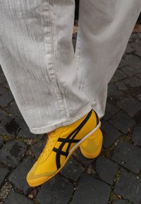 Geel sportieve schoenen met zwarte accenten, suedekappen en een gemusterde zool. Gedragen met lichtgekleurde, gestructureerde broeken op een kasseienvloer.