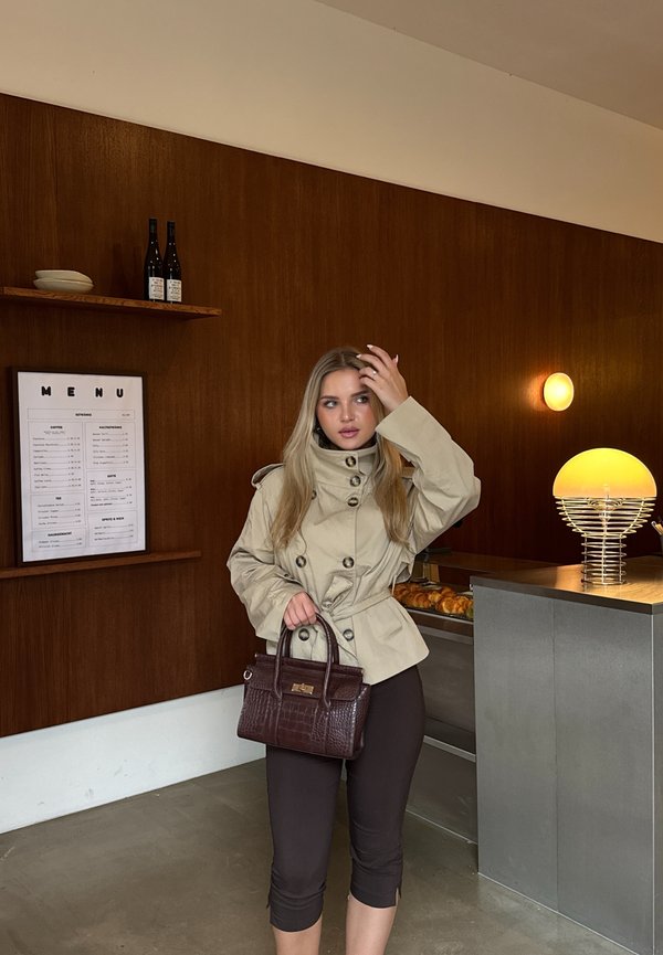 Mujer con chaqueta beige y pantalones cortos negros sostiene un bolso oscuro dentro de una cafetería con paredes de madera y luz cálida.