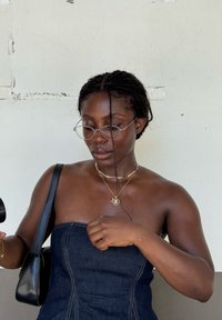 Top in denim senza spalline con cuciture verticali, collane d'oro a strati, occhiali trasparenti rotondi e una borsa nera tenuta in mano contro uno sfondo tenue.